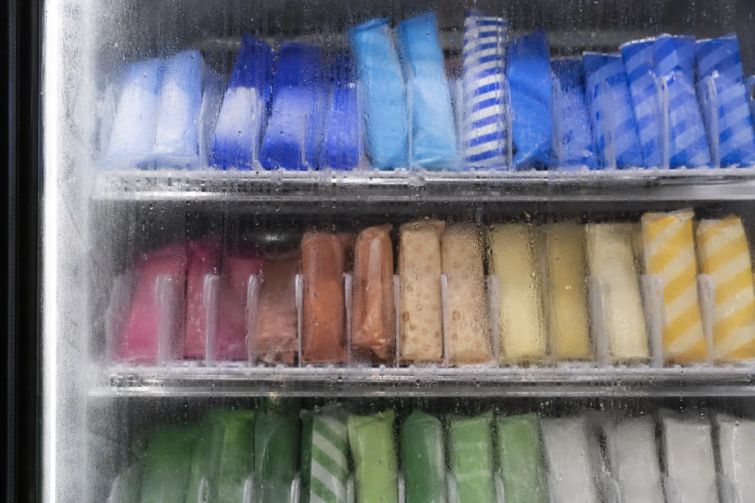 Närbild genom en immig frysruta på rader av enskilt insvepta glassar i olika färger, utan synlig text eller logotyp på förpackningarna